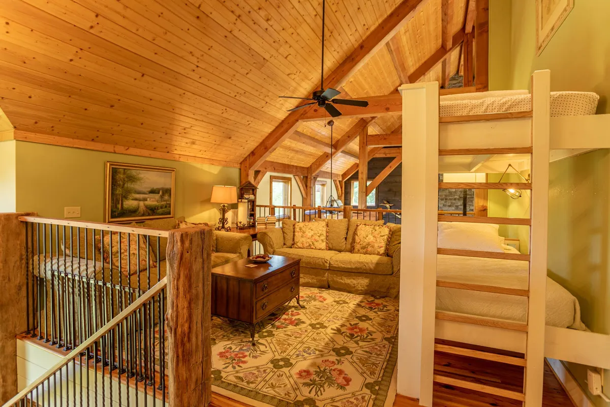 A multi-functional loft area featuring a floral-upholstered sofa, wooden coffee table, and visible bunk bed. The ceiling beams add rustic charm, while the seating invites relaxation or gatherings, reflecting the versatility and coziness described.