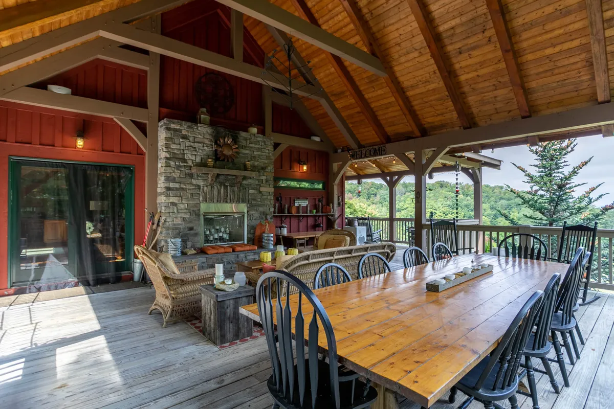 A cozy dining and seating area anchored by a large wooden table, stone fireplace, and comfortable chairs. Floor-to-ceiling windows frame lush green views of the mountains, allowing the beauty of the outdoors to flow inside. The wooden beams and rustic finishes reinforce the theme of mountain luxury blended with natural surroundings.