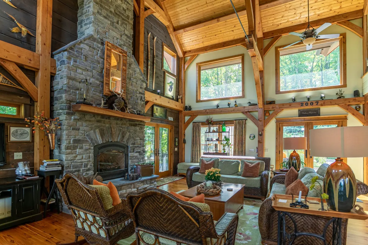 This image captures the great room's soaring vaulted wooden ceiling, a large stone fireplace as the centerpiece, expansive windows with sunlight streaming in, and a cozy seating area arranged for conversation and relaxation. The mountain lodge aesthetic is evident through rustic woodwork and warm decor.