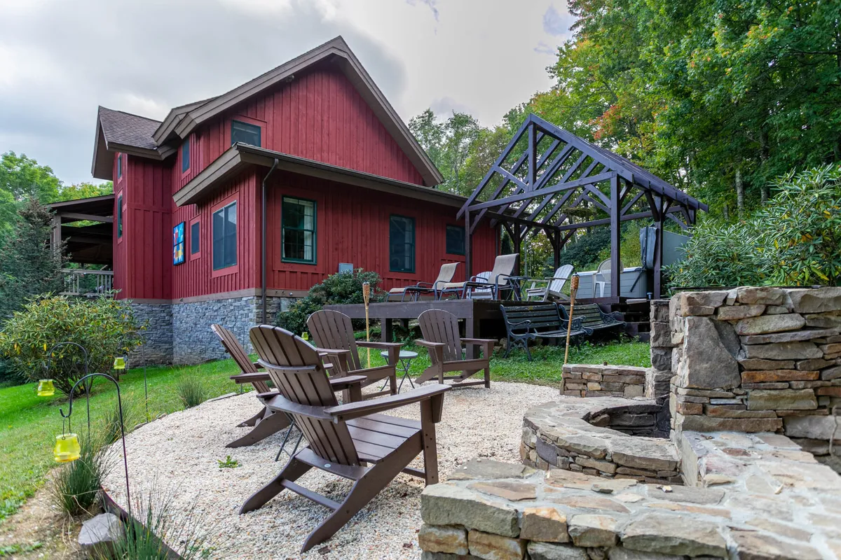 A cozy outdoor gathering space features a stone fire pit encircled by Adirondack chairs, a covered patio, and the striking red exterior of the house. The area is set for relaxation, storytelling, and sharing s'mores under the sky.