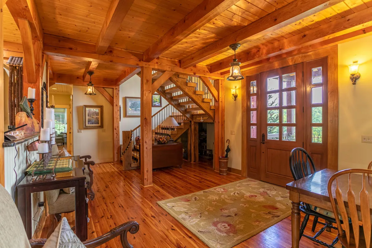 This image captures the entryway of the cabin, featuring warm wooden beams, a welcoming entrance door, a staircase, and a dining table, all awash in inviting light. The scene radiates a sense of arrival and connection, hinting at the seamless transition between shared spaces and private moments.