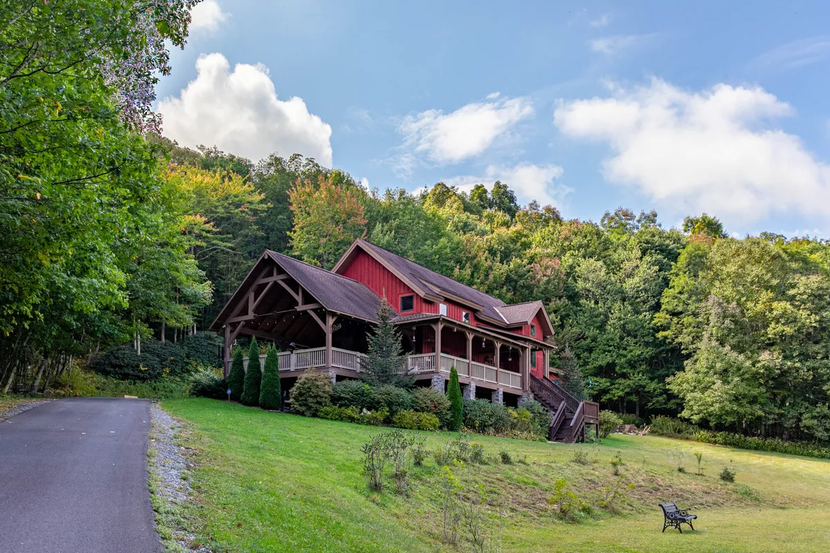 A peaceful exterior shot of the red house nestled among tall, mature trees, with a stone pathway leading to the front porch and a bench in the yard. The lush green surroundings create a sense of seclusion and connection to nature, ideal for outdoor relaxation after a day of adventure.