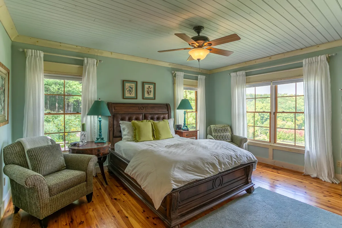 A serene bedroom scene featuring a comfortable bed positioned by large windows, an inviting armchair, a nightstand with a lamp, and tranquil natural light streaming in. The setting is uncluttered, peaceful, and showcases cozy details perfect for restful nights.