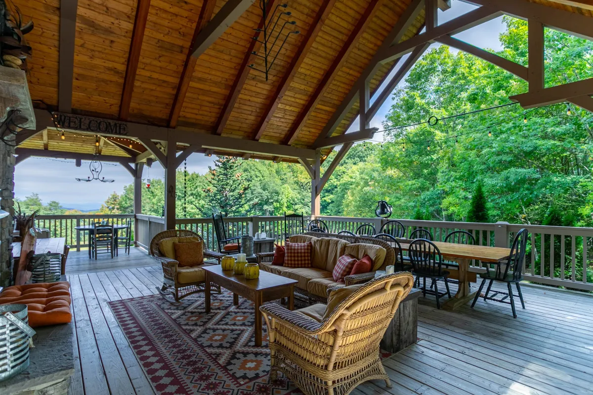 A spacious exterior seating and dining area with comfortable outdoor furniture, a decorative rug, wooden beams overhead, and lush greenery all around. The space invites gatherings, quiet moments in nature, and leisurely meals surrounded by the outdoors.