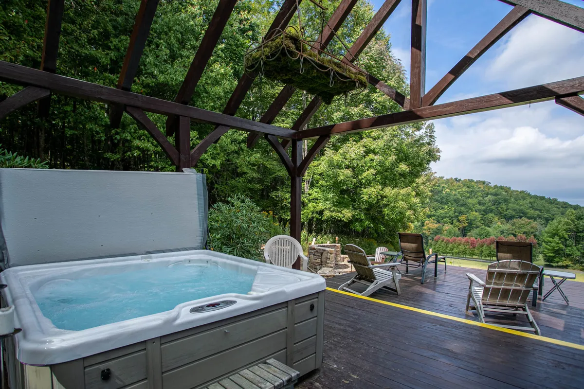 An outdoor hot tub set against a backdrop of wooden beams and lush green landscape, with lounge chairs nearby for relaxation. The area is positioned to maximize privacy and connection to the surrounding scenery, inviting guests to soak while admiring the mountains or stargazing at night.