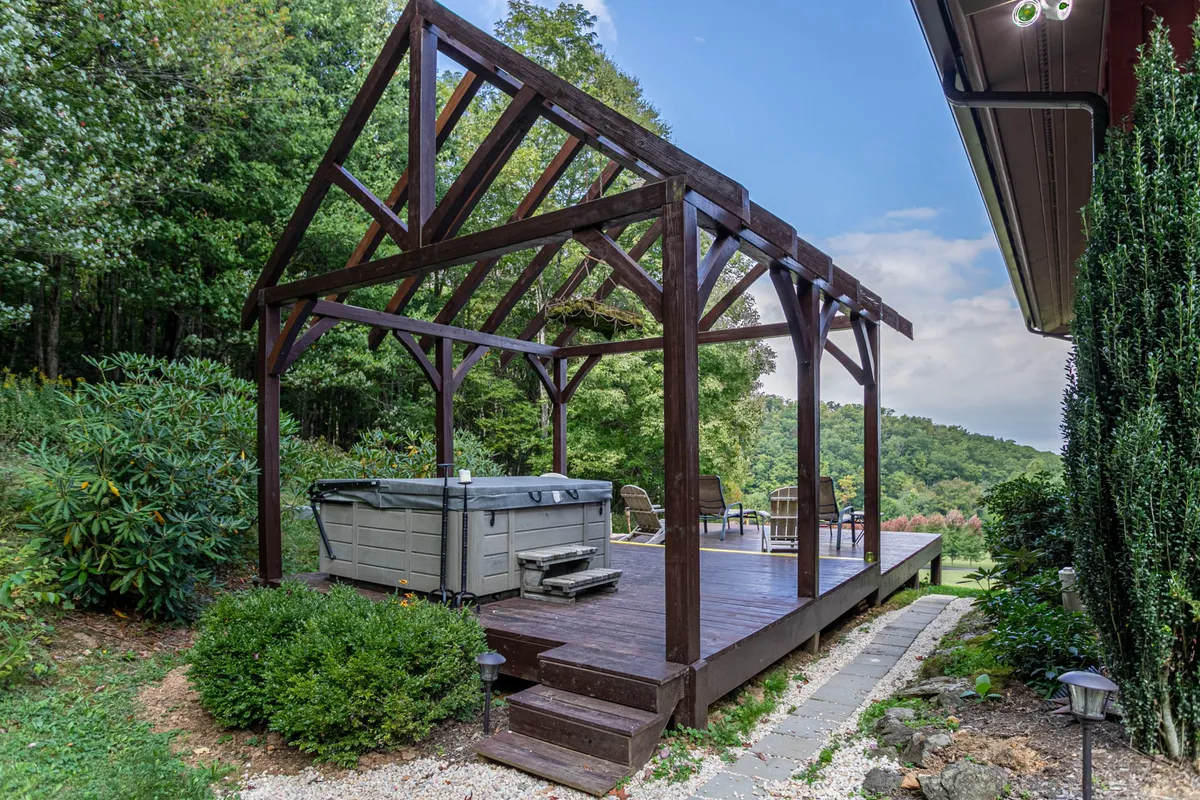 A spacious outdoor deck featuring a wooden gazebo structure, a bubbling hot tub, and surrounding greenery. The inviting setup is ideal for relaxing in the hot tub while taking in the mountain air, with plenty of room for groups to gather and enjoy the scenery.