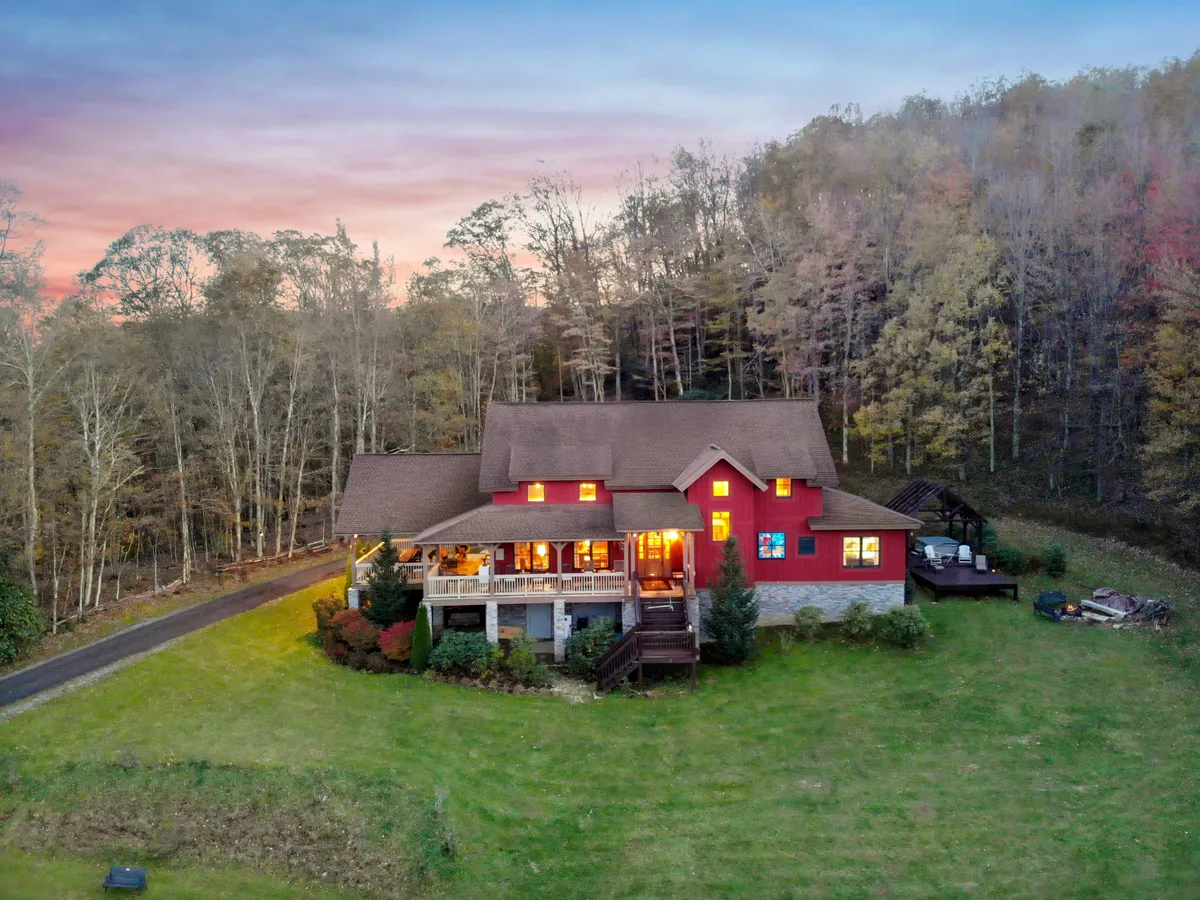 The image showcases a striking red timber-frame cabin nestled among mature trees on a secluded 4-acre property. A spacious deck invites guests to relax and enjoy panoramic mountain views, while the surrounding landscape hints at tranquil sunsets and cozy outdoor gatherings.