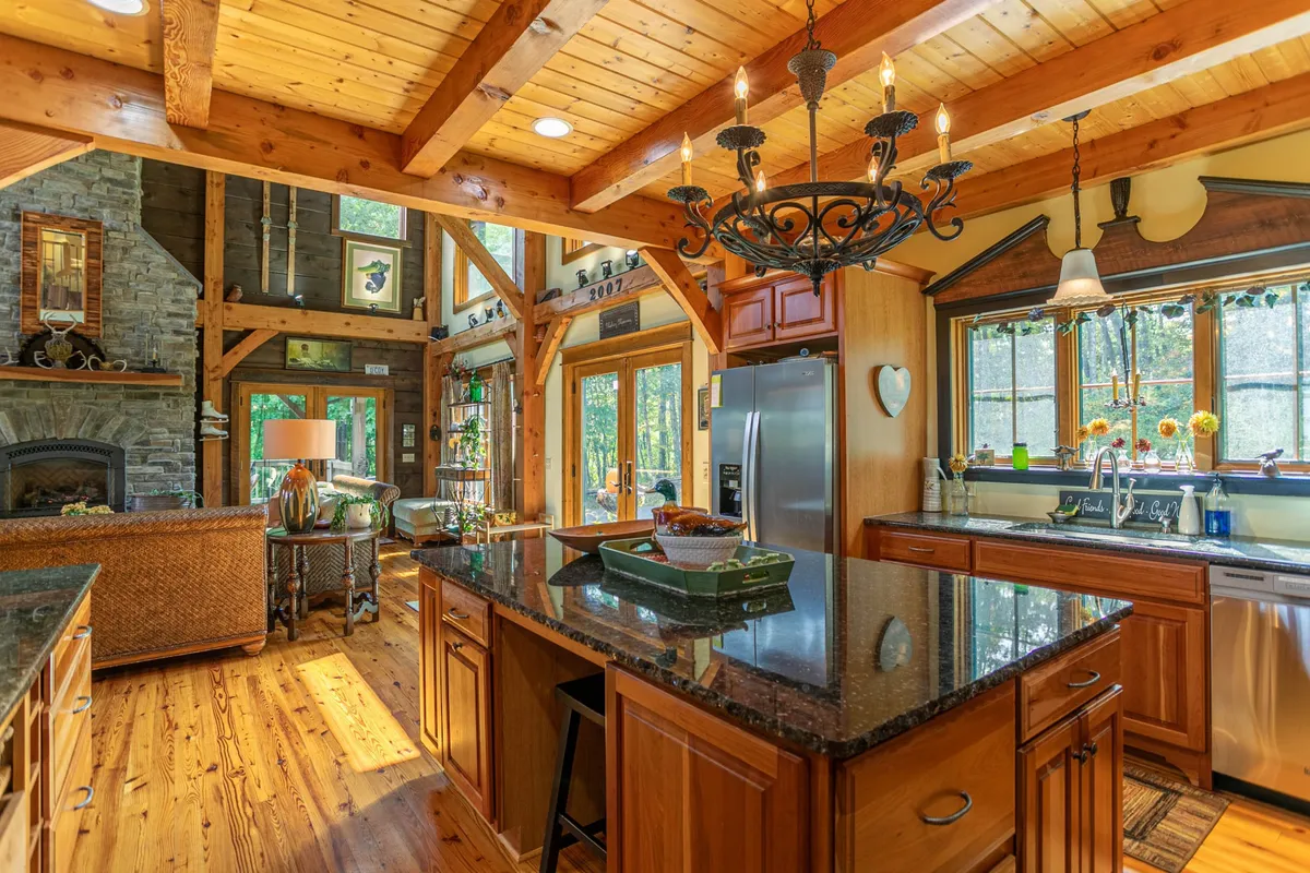 An inviting open-plan kitchen and living area with a central island topped in dark granite, rustic wooden beams and ceiling, large windows framing outdoor views, and a stone fireplace anchoring the space. The layout encourages group interaction and shared cooking experiences.