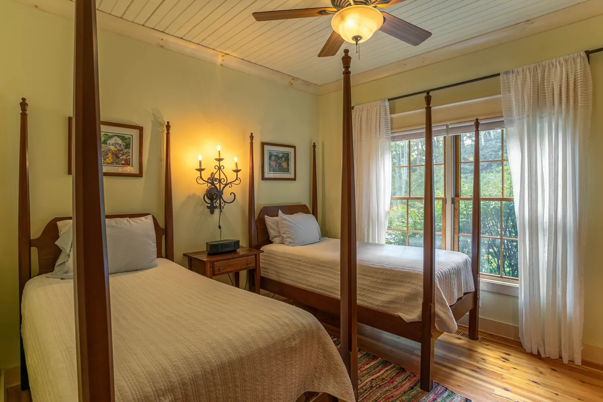 A spacious bedroom with two elegant four-poster beds, a window flooding the room with natural light, an ornate chandelier, and decorative artwork. The room exudes comfort and is ideal for multiple guests or children, aligning with flexible sleeping arrangements.